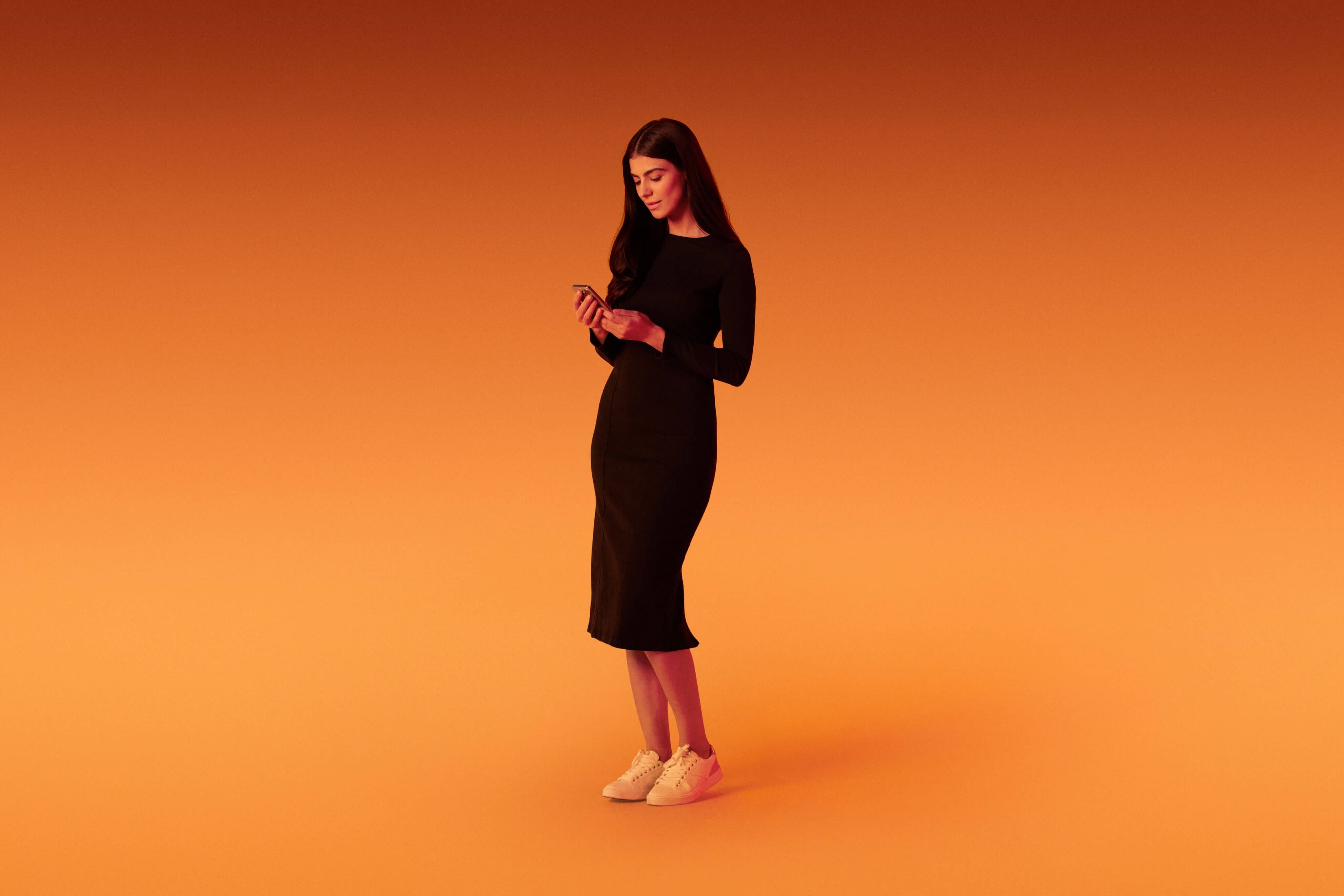 Standing woman engaging with the Xapo Bank app on her mobile phone, overlaid with an orange hue.